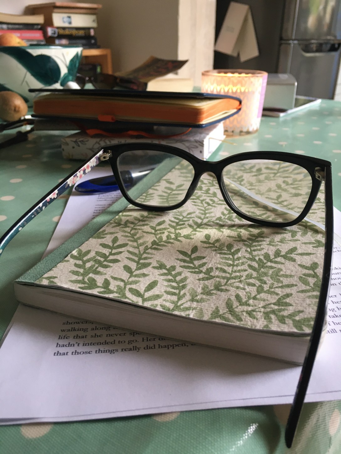 The image is a notebook with a pair of spectacles on top. A writing pen is nearby. In the near distance are more notebooks.