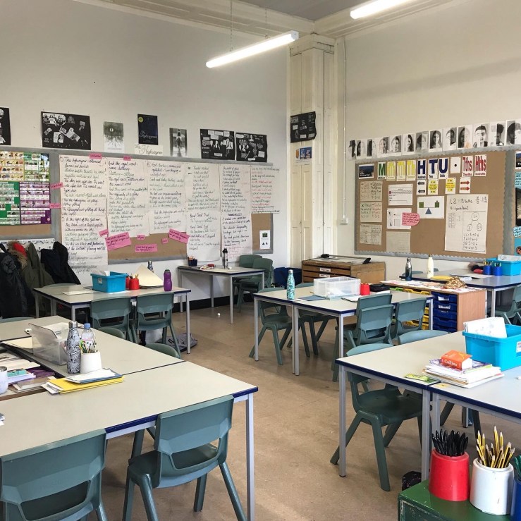 The image is of an empty classroom, walls covered in posters and children's drawings, pens and paper on empty desks with chairs under them. 