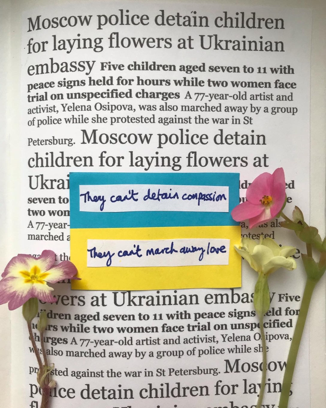 The image is of a news report that children were detained in Moscow for laying flowers at the Ukraine embassy, and a 77 year old woman was marched away in St Petersburg for holding a sign protesting the invasion of Ukraine. Laid on top of the news text are the handwritten words "They can't detain compassion" and "They can't march away love".