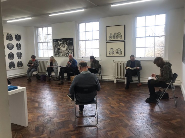 The image is of a group of people sitting in a loose circle in an art gallery, socially distanced from each other, writing in notebooks on their laps.
