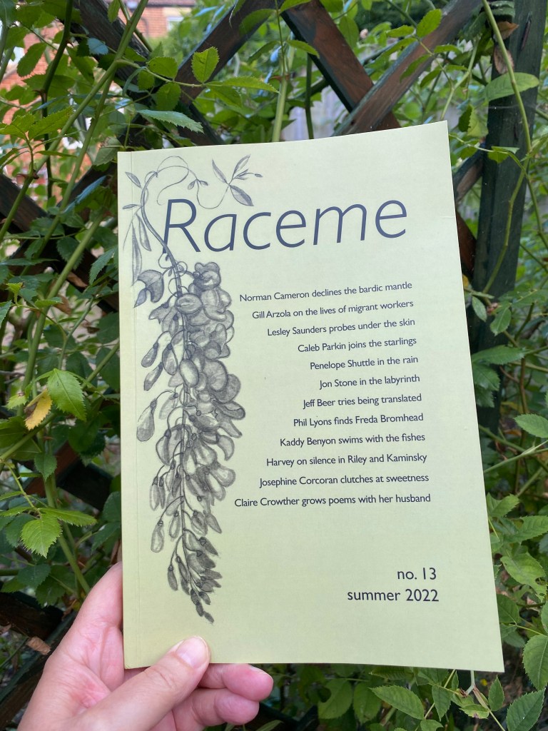 The image is of a magazine being held up by a white hand in front of tangled green shrub growing up a trellis