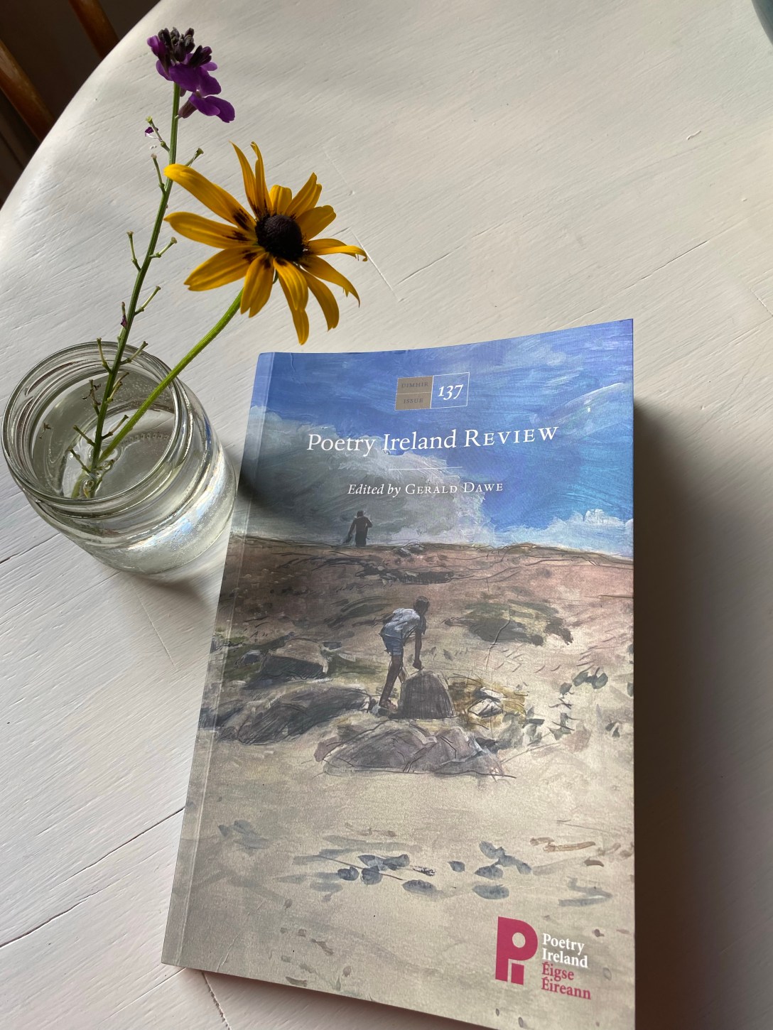 The image is of issue 137 of Poetry Ireland Review which features a painting of two blurred figures clambering up the side of a hill or sand dune beneath a vividly blue, clouded sky